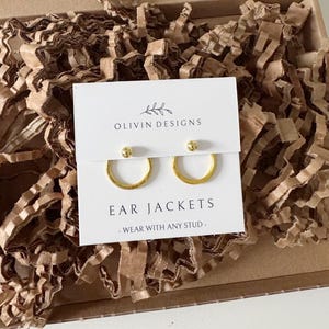 May include: A cardboard box filled with brown shredded paper, containing a white card with the text "OLIVIN DESIGNS" and a pair of gold hoop earrings on a white card that reads "EAR JACKETS - WEAR WITH ANY STUD". A small thank you card is also visible.