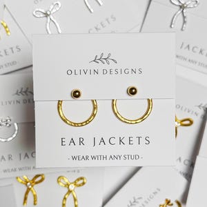 May include: An assortment of earring sets and ear jackets displayed on white cards. The earrings feature designs like bows, hoops, and abstract lines, in gold and silver. The cards are printed with "OLIVIN DESIGNS" and "EAR JACKETS - WEAR WITH ANY STUD."