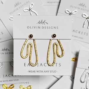 May include: A collection of earrings on white cards. The earrings are in gold and silver tones, featuring bow and abstract designs. The cards display the text "OLIVIN DESIGNS" and "EAR JACKETS - WEAR WITH ANY STUD."