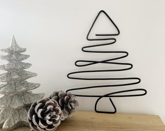 May include: A black wire Christmas tree ornament with a simple, abstract design. The ornament is displayed on a wooden surface with two small, brown pinecones.