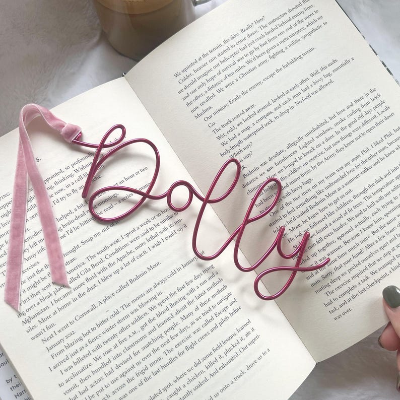 May include: A red metal bookmark shaped like the word "Dolly" is shown on top of an open book. The bookmark is made of thin wire and has a delicate, cursive style.