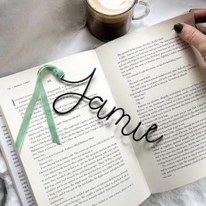 May include: A black metal bookmark shaped like the name "Jamie" is placed on an open book. The bookmark is resting on a green and white ribbon bookmark. The book is open to a page with text about a military experience.