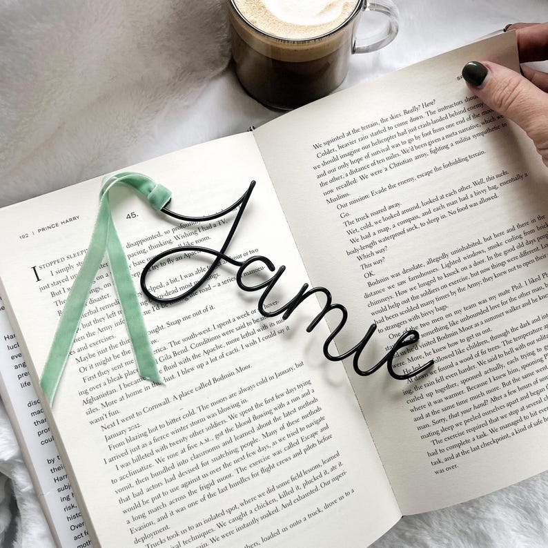 May include: A black metal bookmark shaped like the name "Jamie" is placed on an open book. The bookmark is resting on a green and white ribbon bookmark. The book is open to a page with text about a military experience.