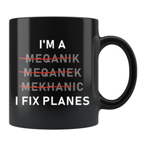 May include: Black ceramic mug with the text "I'M A MEQANIK MEQANEK MEKHANIC I FIX PLANES" in white. The words "MEQANIK", "MEQANEK", and "MEKHANIC" are crossed out in red. The mug has a black handle.