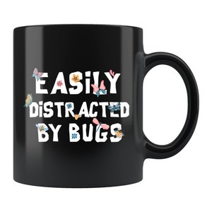 May include: Black ceramic coffee mug with white text that reads "Easily Distracted By Bugs". The text is surrounded by colorful butterflies, dragonflies, and bees.