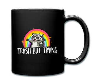 May include: Black coffee mug featuring a cartoon raccoon in front of a rainbow with the text "Trash But Trying".