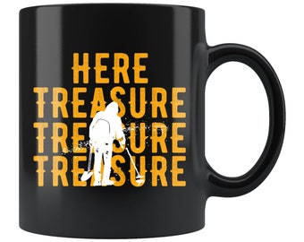Può includere: Tazza da caffè in ceramica nera con una silhouette bianca di una persona che tiene un metal detector. Il testo "HERE TREASURE TREASURE TREASURE" è stampato in giallo sulla tazza.