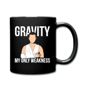 May include: Black coffee mug with white text that reads "GRAVITY MY ONLY WEAKNESS". A cartoon illustration of a person with a broken arm is in the center of the mug.