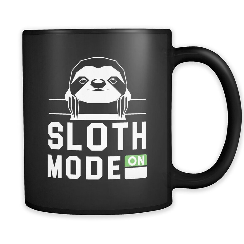 Puede incluir: Taza de caf&eacute; negra con un perezoso blanco asom&aacute;ndose por el borde y el texto "Sloth Mode ON" en letras blancas.