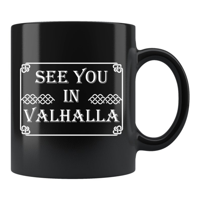 May include: Black ceramic mug with a white rectangular design. The design features the text "SEE YOU IN VALHALLA" in a bold, serif font, framed by decorative elements. The mug has a curved handle.