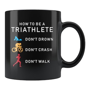 Könnte beinhalten: Schwarze Kaffeetasse mit weißem Text, der "How to be a Triathlete" lautet, mit drei Symbolen und Text unter jedem Symbol: "Don't Drown", "Don't Crash" und "Don't Walk".
