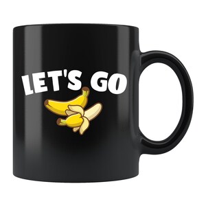 Puede incluir: Taza de café negra con un diseño blanco que dice "Let's Go" con una imagen de dibujos animados de dos plátanos.
