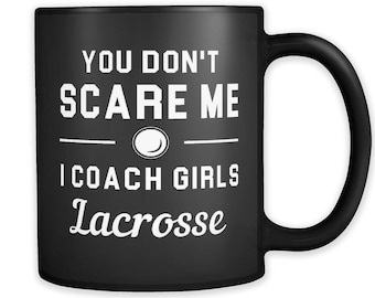 May include: Black ceramic mug with the text "YOU DON'T SCARE ME I COACH GIRLS Lacrosse" in white. The mug features a lacrosse ball graphic. The mug has a black handle and a glossy finish.