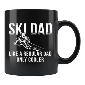Könnte beinhalten: Schwarze Keramik-Tasse mit weißem Aufdruck „SKI DAD“ und einer Skifahrer-Grafik. Darunter steht „LIKE A REGULAR DAD ONLY COOLER“. Die Tasse hat einen gebogenen Henkel.