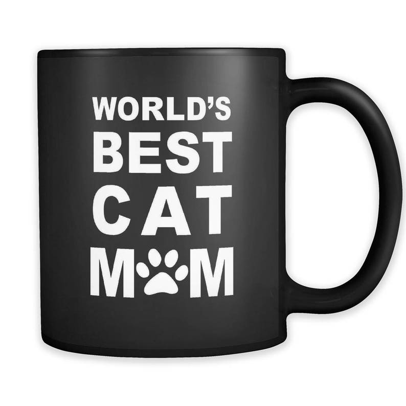Puede incluir: Taza de caf&eacute; de cer&aacute;mica negra con texto blanco que dice "World's Best Cat Mom" con una huella de pata en lugar de la "O".