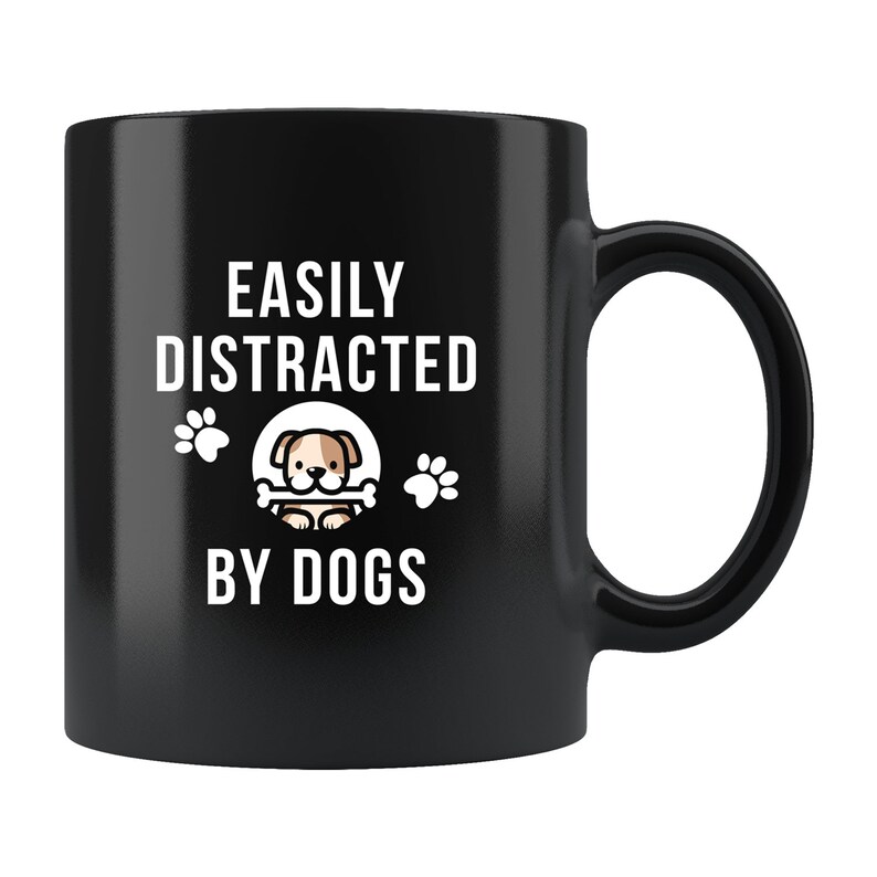 May include: Black ceramic coffee mug with white text that reads "Easily Distracted By Dogs". A cartoon dog with a bone is in the center of the mug.