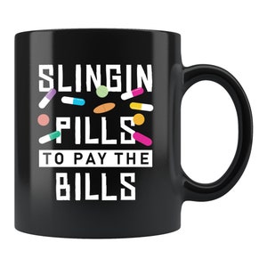 Puede incluir: Taza de café negra con texto blanco que dice "Slingin Pills To Pay The Bills" con píldoras de colores esparcidas alrededor del texto.