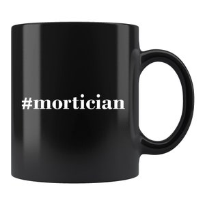 May include: Black ceramic coffee mug with a white hashtag and the word "mortician" printed on the front.