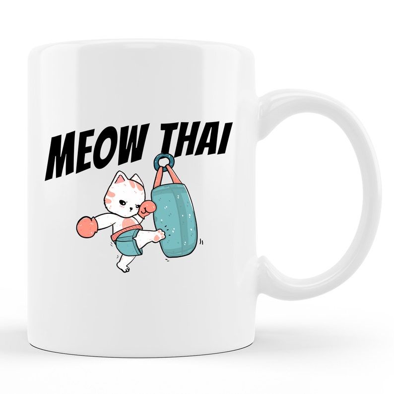 Peut inclure: Une tasse en c&eacute;ramique blanche avec une illustration de dessin anim&eacute; d'un chat qui donne un coup de pied dans un sac de frappe bleu. Le texte "MEOW THAI" est imprim&eacute; en noir au-dessus de l'illustration.