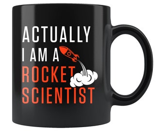 Może przedstawiać: Czarny kubek do kawy z białym tekstem, który brzmi "Actually I am a Rocket Scientist" z grafiką czerwonej rakiety i białą chmurą dymu.