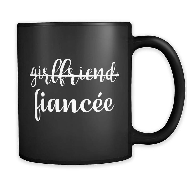 May include: Black coffee mug with white text that reads "girlfriend" with a line through it and "fianc&eacute;e" below it.