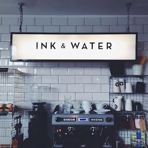 Puede incluir: Un letrero en blanco y negro con el texto "INK & WATER" colgado del techo sobre un mostrador de cafetería. El mostrador tiene una máquina de espresso plateada y un frasco de vidrio con malvaviscos blancos.