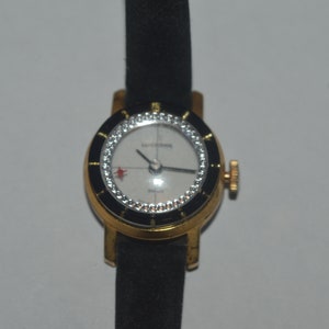 May include: A gold-tone wristwatch with a black leather band. The watch face is white with black Roman numeral hour markers and a black second hand. The watch face is surrounded by a ring of clear crystals.
