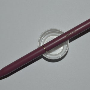 May include: A burgundy pen with gold accents and the word "BILLIE" printed on the side. The pen is resting on a white plastic lid. The pen has a gold tip and a gold band near the top.