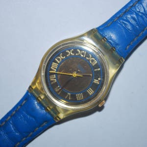 Puede incluir: Un reloj de pulsera dorado con una correa de cuero azul. La esfera del reloj tiene marcas de números romanos y un fondo negro.