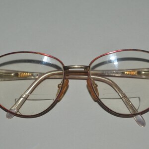 May include: A pair of gold-rimmed glasses with red accents. The glasses have a rounded, oval shape and clear lenses.