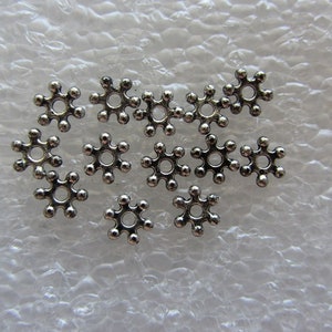 May include: A group of 14 silver snowflake-shaped metal beads. The beads have a small hole in the center and are designed to be used for jewelry making.