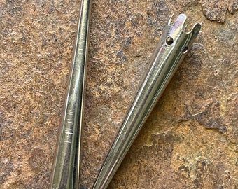 May include: Two silver metal tools with a tapered design. One tool has a rounded ball at the end, while the other has a forked end with small holes. The tools are on a brown, textured surface.