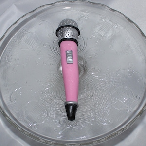 May include: A pink and silver microphone cake topper. The microphone is decorated with a silver grill and a black button.
