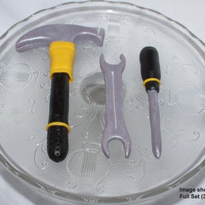 May include: A set of three tools made of fondant, including a hammer, wrench, and screwdriver. The tools are gray with yellow accents.