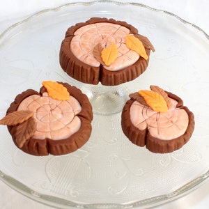May include: Three brown and orange fondant cake toppers shaped like tree stumps with leaves. The toppers are on a clear glass cake stand.