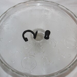 May include: A clear glass cake stand with a decorative pattern. The stand has two black metal hooks on the top.