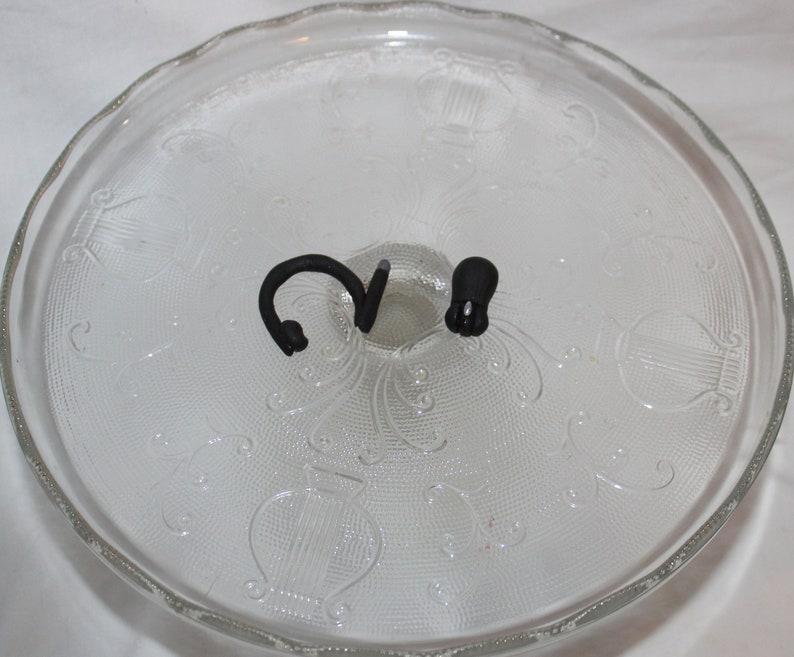 May include: A clear glass cake stand with a decorative pattern. The stand has two black metal hooks on the top.