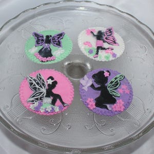 May include: Four round, decorated sugar cookies with a fairy silhouette design. Each cookie features a different color background: green, pink, purple, and white. The cookies are arranged on a clear glass plate.