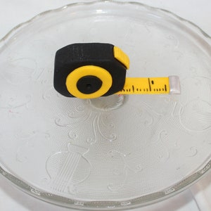 May include: A black and yellow measuring tape with a silver end cap. The tape is on a clear glass plate with a decorative pattern.