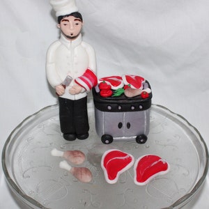 May include: A white fondant chef figurine holding a knife and a red and white striped towel, standing next to a gray fondant grill with red fondant steaks, tomatoes, and green fondant lettuce. There are also two fondant chicken legs and two fondant steaks on a clear glass cake stand.