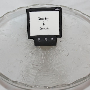May include: A black and white cake decoration shaped like a television screen with the text "Darby & Shane" on the screen.