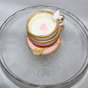 May include: A decorative teacup and saucer set with gold trim, a pink heart, and a gold spoon. The teacup is white and pink, and the saucer is pink. The set is on a clear glass plate.