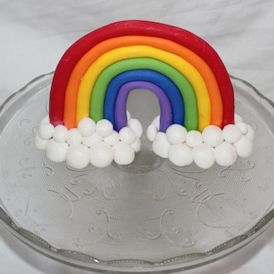 May include: A rainbow cake topper made of fondant, with a red, orange, yellow, green, blue, and purple rainbow arch. The rainbow is sitting on a bed of white fondant clouds.