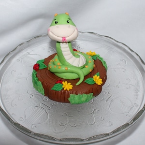 May include: A green snake cake topper with a pink tongue, sitting on a brown tree stump with green leaves and yellow flowers.