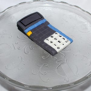 May include: A gray, blue, and yellow calculator-shaped confection sits on a clear glass plate with a decorative pattern. The calculator has white number keys and a black display screen.