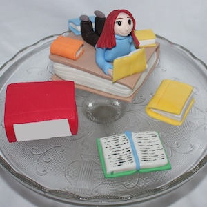 May include: A cake decorated with a figurine of a person reading a book, surrounded by miniature books. The cake is on a clear glass cake stand.