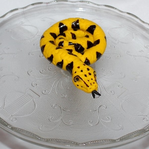 May include: A yellow and black snake figurine coiled on a white cake stand with a decorative pattern.