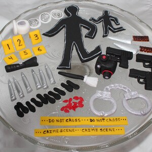 May include: A white cake decorated with a crime scene theme. The cake features a black silhouette of a person, a flashlight, bullets, handcuffs, a knife, and a gun. The cake also has yellow numbers and the text "DO NOT CROSS... DO NOT CROSS... ... CRIME SCENE...CRIME SCENE..."
