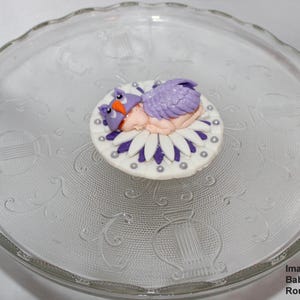 May include: A purple and white fondant baby topper with a baby sleeping on a flower. The topper is on a round glass cake stand.