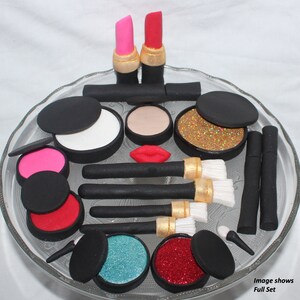 May include: A set of fake makeup cake toppers, including lipsticks, eyeshadow palettes, brushes, and glitter. The image shows a full set of cake toppers.